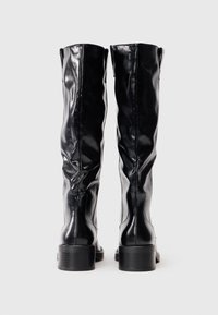Botas largas hasta la rodilla de color negro con un acabado de charol brillante, puntera redondeada y tacón bajo. Costuras decorativas recorren los laterales.