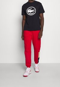 Mężczyzna w czarnej koszulce Lacoste, czerwonych dresowych spodniach z beżowymi i granatowymi łatami oraz białych-czerwonych sneakersach stoi przed jednolitą ścianą.