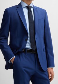 Niebieska marynarka garniturowa z wyciętymi klapami, dwoma guzikami i pojedynczą kieszenią na piersi. W połączeniu z jasnoniebieską koszulą i ciemnym krawatem. Gładka tekstura.