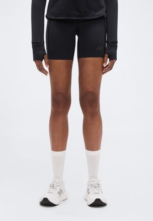 Pantalones cortos negros ajustados con una textura suave, combinados con calcetines blancos y zapatillas claras. El conjunto se complementa con una camiseta negra de manga larga.