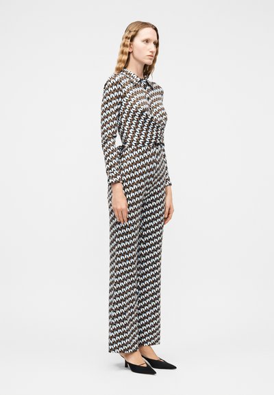 Γυναίκα με μακριά κυματιστά μαλλιά, που φοράει ολόσωμη φόρμα με μακριά μανίκια σε καφέ, λευκό και μπλε σχέδιο και μαύρες μυτερές γόβες, στέκεται προφίλ.