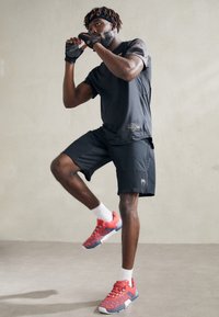 Schwarzes kurzärmliges Sportshirt, schwarze Shorts, rot-blaue Sportschuhe und Fitnesshandschuhe. Das Outfit zeichnet sich durch verschiedene Texturen und ein sportliches Design aus.