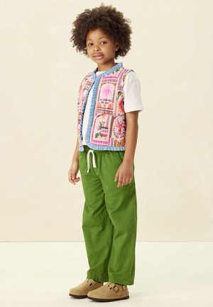 Kleurige gewatteerde vest met bloempatronen over een lichtblauw shirt, gecombineerd met groene broek en beige klompen.