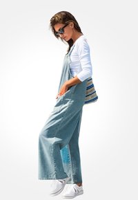 Dżinsowe ogrodniczki z szerokimi nogawkami w jasnoniebieskim kolorze, w połączeniu z białą koszulą z długim rękawem i białymi trampkami, z wzorzystą torbą.