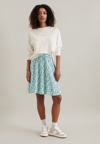 Sweatshirt blanc avec un logo subtil, associé à une jupe bleu clair avec un motif noir et vert éparpillé, portée avec des baskets.