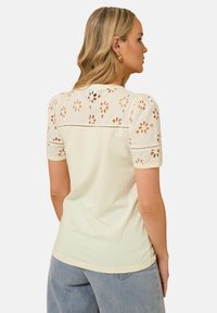Cream-colored T-shirt with a floral cut-out design on the back, short puff sleeves, and a soft textured finish.
