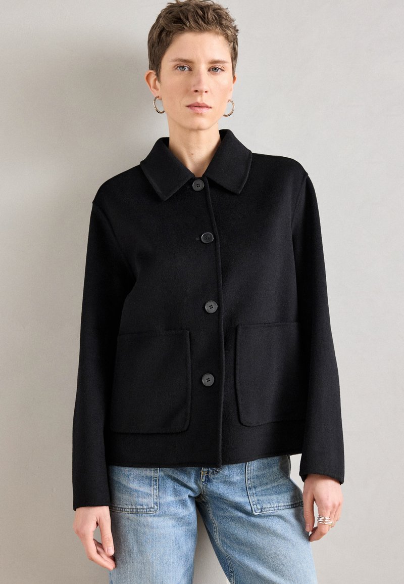 Blazer preto feito à medida com gola de colarinho, botões frontais e dois grandes bolsos de remendo. Confeccionado em tecido texturizado. Combinado com jeans azul claro.