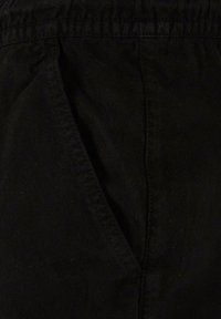 Gros plan sur un pantalon en tissu noir montrant une poche latérale avec des coutures visibles et une ceinture élastique.