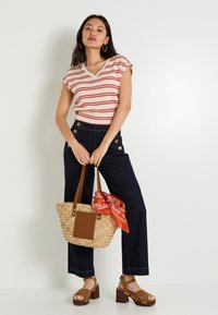 Top de punto a rayas en crema y rojo, pantalones de mezclilla oscuros de pierna ancha con detalles de botones, bolso de paja con asa marrón y bandana naranja, tacones marrones.