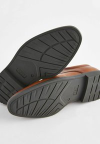 Semelles de chaussures en cuir marron avec une semelle extérieure en caoutchouc noir texturé, présentant un motif d'adhérence et le nom de la marque "next" estampé.