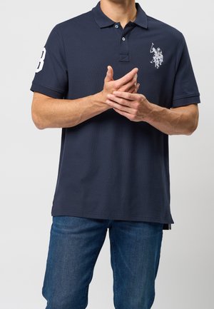 Marineblaues Polohemd mit strukturierter Stoffqualität, versehen mit einem weiß gestickten Logo auf der Brust und einer Zahl auf dem Ärmel.