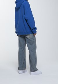 Felpa blu con cappuccio e vestibilità rilassata, abbinata a jeans di denim azzurri e sneakers bianche, vista di spalle.
