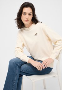 Felpa color crema con collo rotondo a coste, maniche lunghe e un piccolo ricamo di croissant marrone sul petto. Indossata con jeans blu.