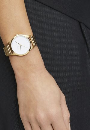Gouden metalen polshorloge met een witte wijzerplaat, drie wijzers en een brede geschakelde band. Achtergrond met een donkere, gestructureerde stof.