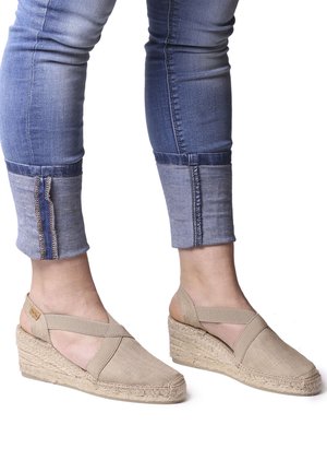 Beige canvas espadrille wedge sandals with cross-straps, woven jute detailing, and a textured rubber sole. Worn with cropped denim jeans.