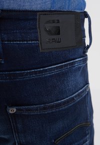 Ciemnoniebieskie dżinsy z denimu z czarną skórzaną naszywką z napisem "G RAW" nad tylną kieszenią oraz widocznymi przeszyciami.