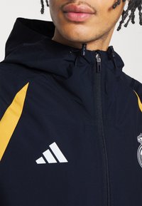 Man draagt een marineblauwe capuchonjas met gele panelen, wit Adidas-logo en een deels zichtbare Real Madrid-wapen op de borst.