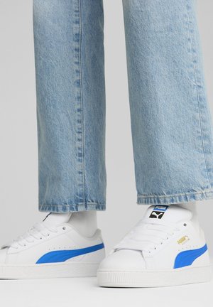 White sneakers with blue accents, featuring a textured surface, rounded toe, and rubber sole. Worn with light blue denim pants.