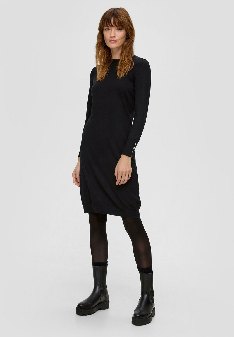 Frau mit glattem rotbraunem Haar, die ein schwarzes langärmliges knielanges Kleid, schwarze Strumpfhosen und schwarze Stiefeletten trägt und vor einem weißen Hintergrund steht.