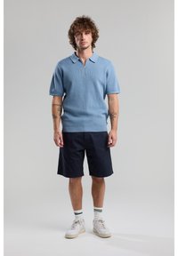Light blue short-sleeve knit polo with zipper, paired with navy shorts. Model wears white sneakers with green striped socks. Smooth texture.