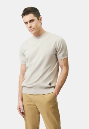 Giovane uomo che indossa una maglia beige a maniche corte in maglia e pantaloni beige, in piedi con una mano in tasca davanti a uno sfondo semplice.