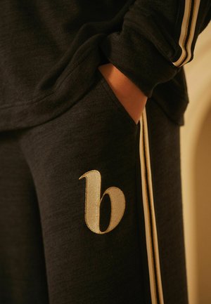 Black sweatpants with a textured fabric, featuring gold side stripes and a decorative embroidered letter "b" on the thigh.