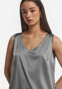 Silvrig satinärmlös topp med V-ringning, slät textur och en lätt lyster; har minimal sömning och en diskret sidficka.