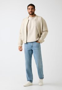 Lichtbeige bomberjack over een crèmewitte sweatshirt, gecombineerd met lichtblauwe spijkerbroek en witte sneakers. Eenvoudig ontwerp met geribbelde manchetten.