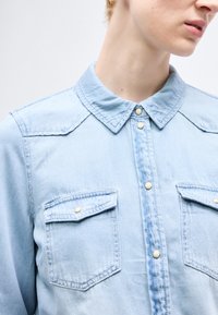 Κοντινό πλάνο ενός ατόμου που φοράει ένα ανοιχτό μπλε πουκάμισο από τζιν με κουμπιά τύπου snap και δύο εμπρόσθιες τσέπες με καπάκι, κοιτάζοντας ελαφρώς προς τα δεξιά.