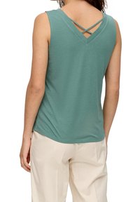 Donna con top senza maniche di colore teal con spalline incrociate sul retro e pantaloni beige chiaro, visibile da dietro.