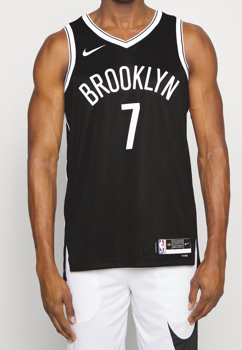 Homme portant un maillot de basket noir de Brooklyn avec le numéro 7 et un short blanc, debout devant un fond uni.