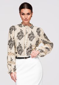 Femme portant un chemisier beige avec des motifs floraux noirs et des détails en dentelle, associé à une jupe blanche et une ceinture noire, posant avec la main sur la hanche.