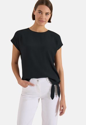 Femme portant un haut noir à manches courtes avec un détail noué sur le côté et un pantalon blanc taille haute, debout avec une main dans la poche.