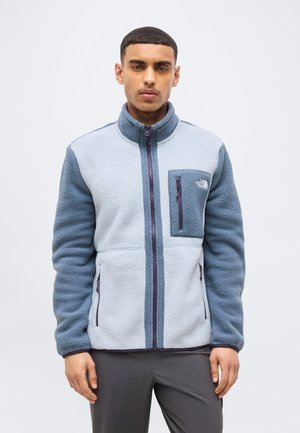 Uomo che indossa una giacca in pile con cerniera color azzurro chiaro e grigio, con tasca sul petto e logo The North Face, in piedi davanti a uno sfondo neutro.