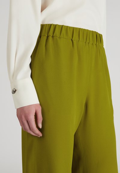 Groene stoffen broeken met een elastische tailleband, gecombineerd met een witte blouse met lange mouwen en een accent bij de manchetten. Gladde textuur.