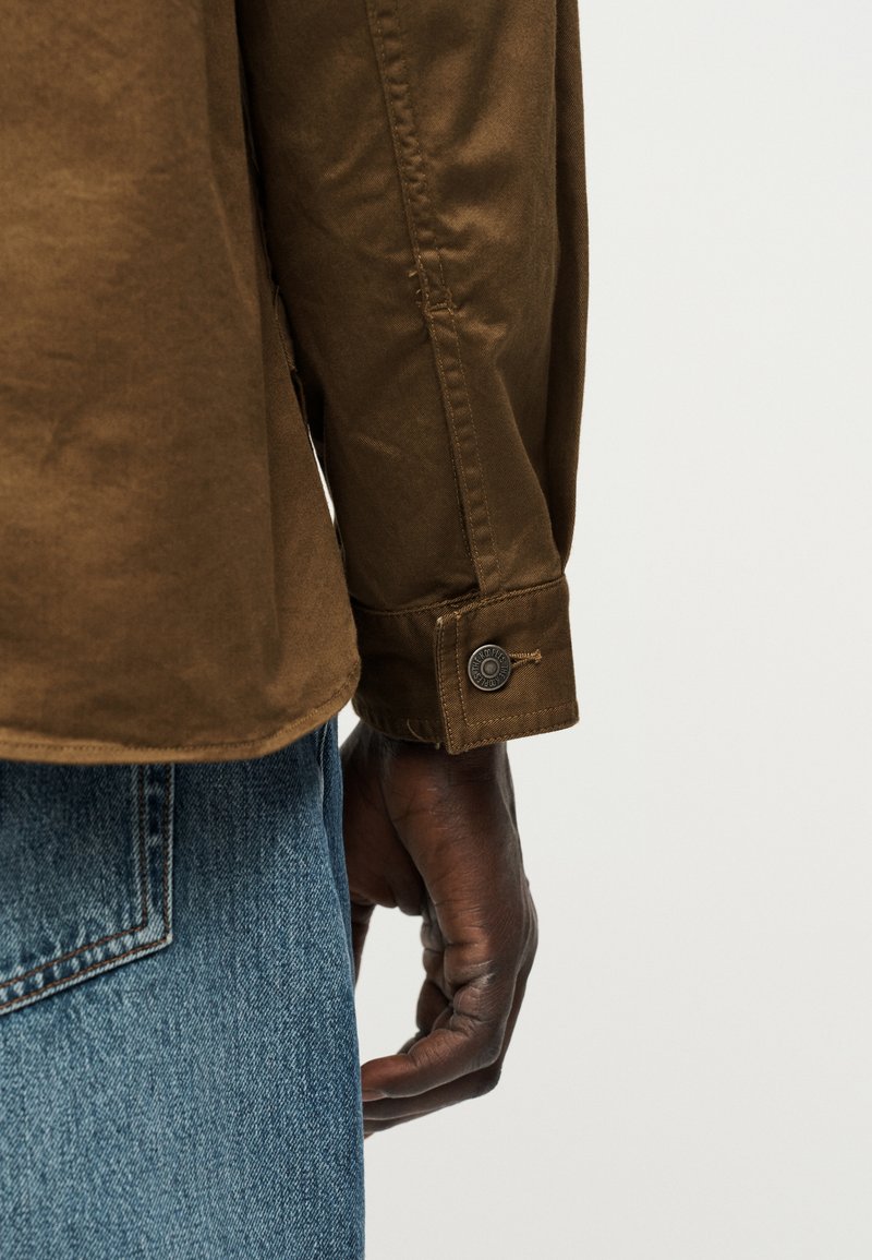 Dunkelhäutige Hand, die ein braunes Langarmhemd mit geknöpftem Manschettenbereich trägt, neben der Tasche einer blauen Denim-Jeans vor einfarbigem Hintergrund.