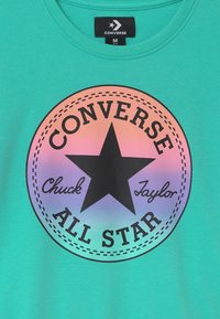 Camiseta verde con un logo circular centrado. El logo tiene un degradado que va del rosa al púrpura, con texto negro y una estrella en el centro.