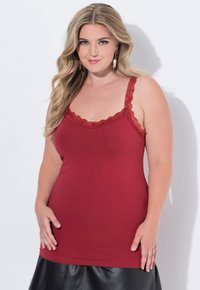 Blonďatá žena s dlouhými vlasy, oblečená v červeném topu s krajkovými detaily a černé sukni, stojící proti bílému pozadí.