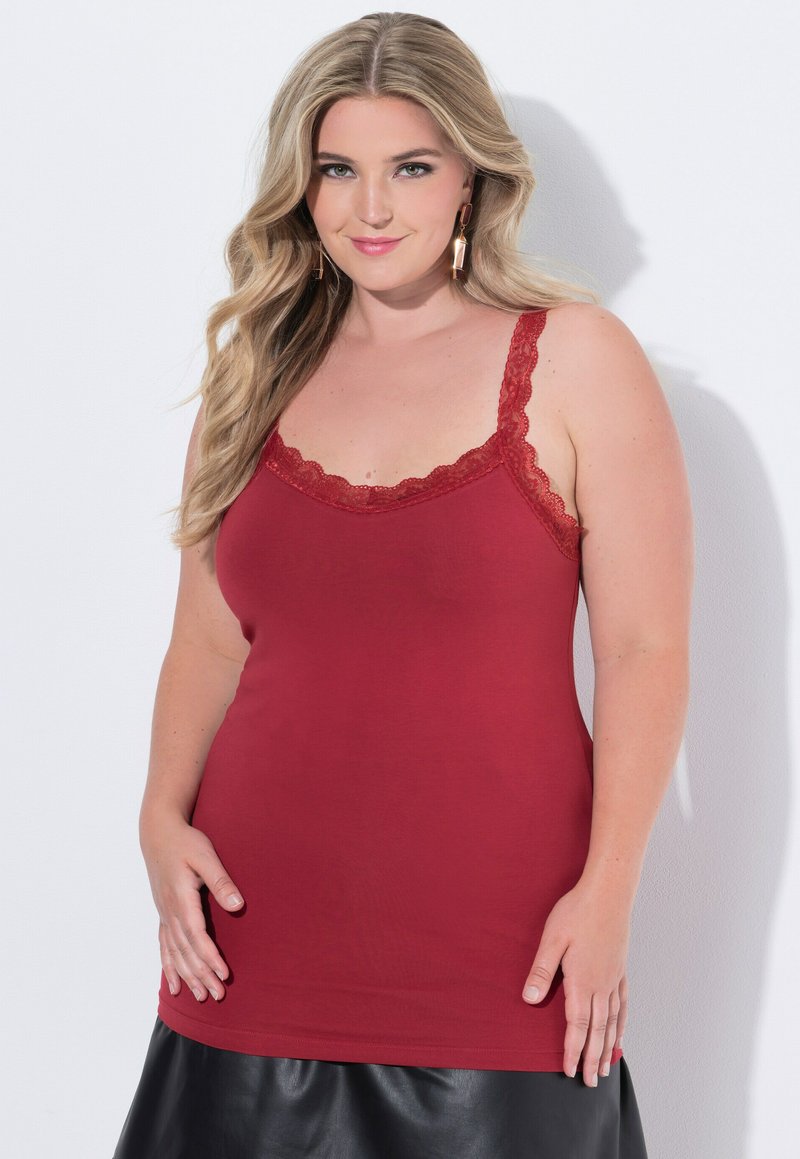 Blonďatá žena s dlouhými vlasy, oblečená v červeném topu s krajkovými detaily a černé sukni, stojící proti bílému pozadí.