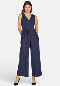 Vrouw staat met handen achter haar rug, gekleed in een mouwloze marineblauwe jumpsuit met een strikceintuur en zwarte hakken, tegen een witte achtergrond.