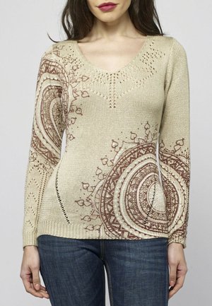 Mujer que lleva un suéter beige de punto con patrones circulares marrones de mandala en la manga y el frente, combinado con jeans azules.
