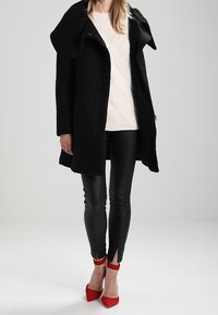 Black textured coat with oversized lapels, worn over a light knit sweater, paired with shiny black leggings and red pointed-toe heels.