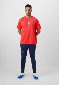 Rød Nike t-shirt med et blåt Paris-emblem, kombineret med marineblå leggings med "PARIS" i guld. Spilleren har hvide fodboldstøvler med blå accenter på.