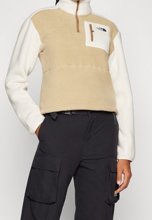 Beige ja valkoinen fleece-pullover, jossa neljänneksen mittainen vetoketju ja rintatasku, yhdistetty mustiin kuorihousuihin, joissa on läppätaskut.