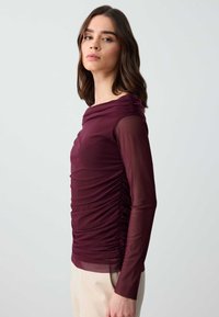 Bordeauxfarbene Bluse mit langen Ärmeln, Raffung im Stoff, durchsichtigen Ärmeln und taillierter Silhouette, kombiniert mit hellen Hosen.