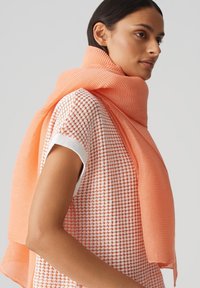 Korallenstreifen-Schal um den Hals drapiert, kombiniert mit einem strukturierten, weißen Oberteil mit Korallenmuster und kurzen, gesäumten Ärmeln.