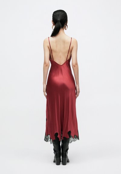 Robe rouge satinée avec un dos en V, des fines bretelles et des détails en dentelle noire à l'ourlet. Le mannequin porte des bottes noires montantes jusqu'aux genoux.