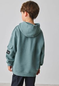 Menta zöld kapucnis pulóver laza szabással, bordázott mandzsettákkal és oldalsó foltzsebbel, fekete címkével díszítve. Puha, texturált anyag.