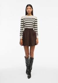 Črtasta bež in črna majica z dolgimi rokavi, rjeno semišasto krilo z visokim pasom in žepi ter črni škornji z nabrušenimi konicami do kolen.