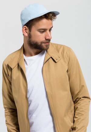 Lys blå caps med rund brem, iført med en lys beige jakke med glidelås. Under er det en hvit t-skjorte. Stoffet er glatt med minimale detaljer.